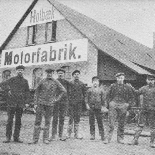 Sort-hvid foto af syv personer foran et hus med teksten Holbæk Motorfabrik.