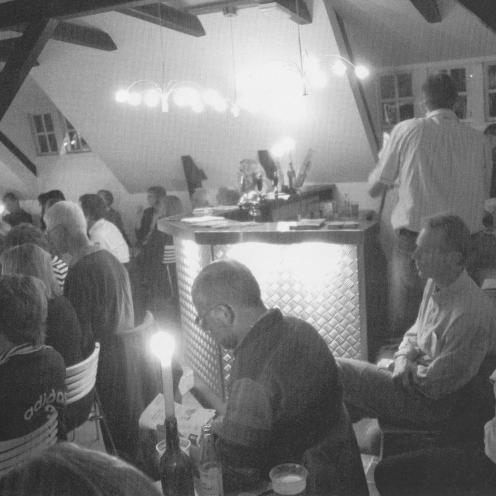 Sort-hvid foto af publikum siddende ved borde med levende lys. Intim stemning og bjælkeloft.