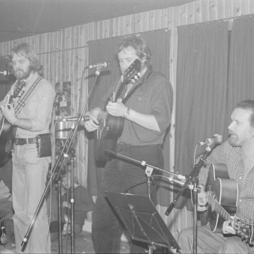Sort-hvid foto af tre musikere, der spiller akustisk guitar og synger, på en lille scene.