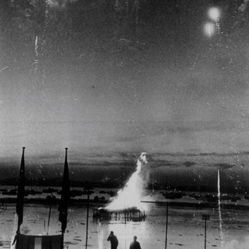 Sort-hvid foto af bål på vandet og fyrværkeri på en mørk himmel.