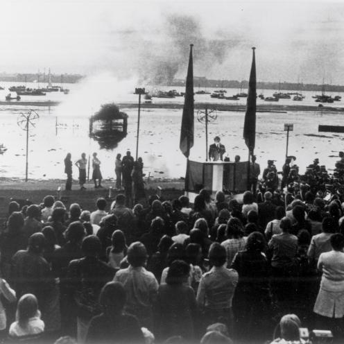 Sort-hvid foto af en menneskemængde foran en talerstol foran fjorden. Der brænder et bål på vandet.