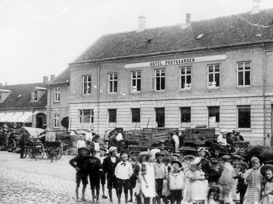 Sort-hvid foto af Hotel Postgården med hestevogne foran og en flok børn forrest i billedet.