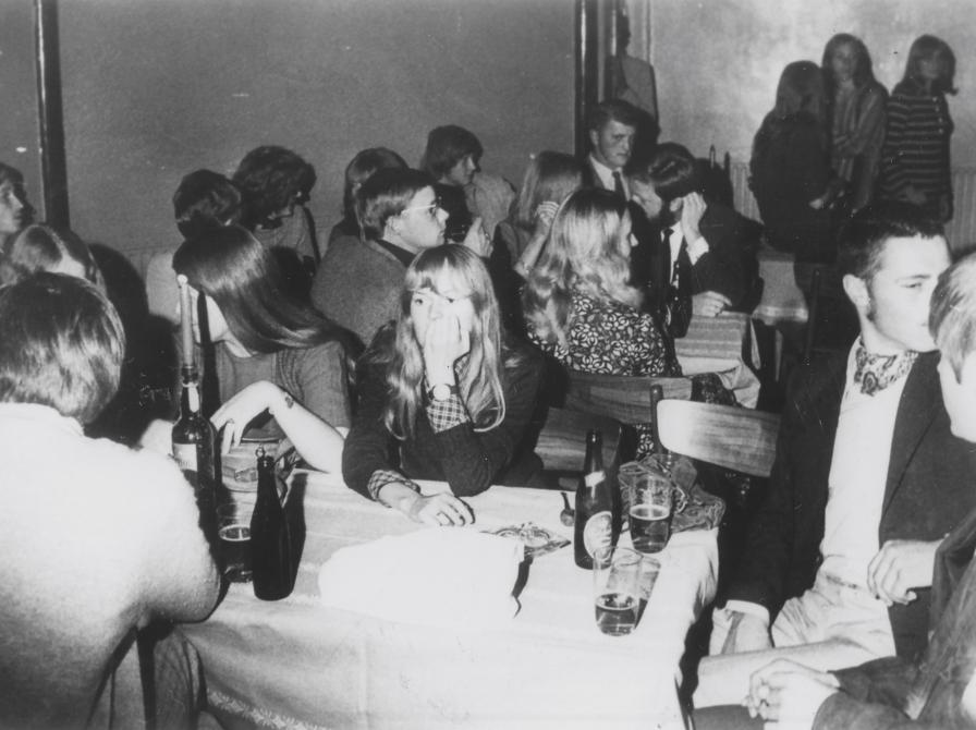 Sort-hvid foto af unge siddende ved et bord med hvid dug og ølflasker.